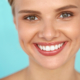 Portrait d'une femme souriante aux yeux verts et aux dents blanches parfaites, posant devant un fond bleu clair.